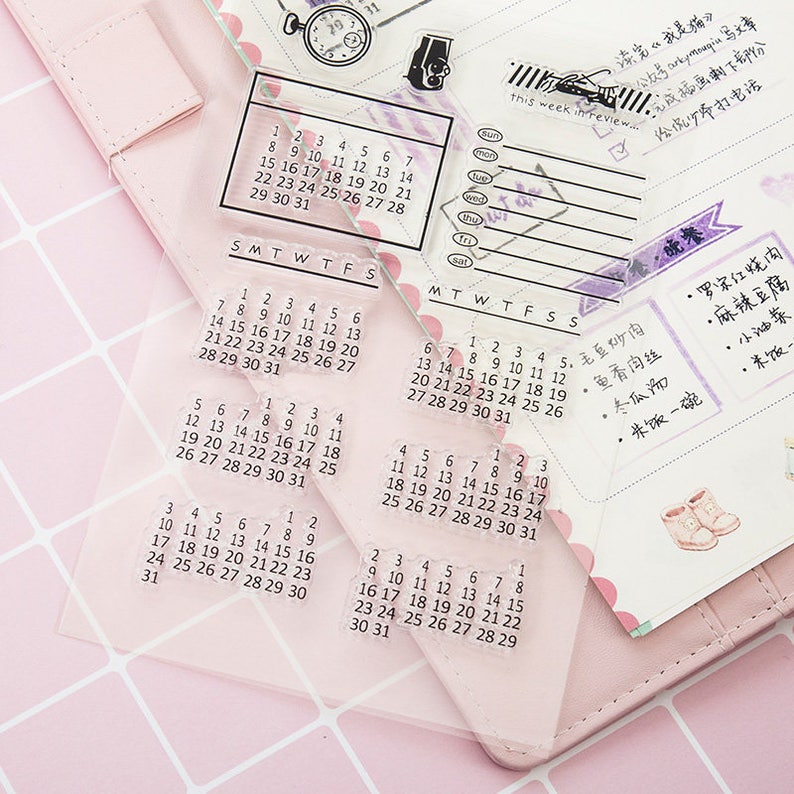 Pu&ograve; includere: Timbri calendario in acrilico trasparente con vari layout mensili e settimanali. I timbri includono numeri, giorni della settimana ed elementi decorativi. I timbri sono su uno sfondo rosa.