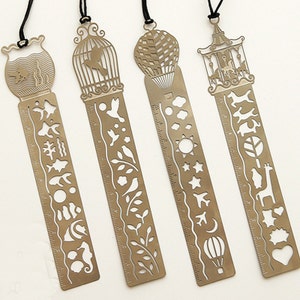 K&ouml;nnte beinhalten: Satz mit vier goldenen Lesezeichen aus Metall mit verschiedenen Designs. Jedes Lesezeichen hat auf der einen Seite ein Lineal und auf der anderen Seite ein dekoratives Design. Die Designs umfassen einen Vogelk&auml;fig, einen Hei&szlig;luftballon, ein Karussell und ein Seepferdchen.