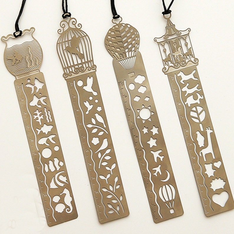 K&ouml;nnte beinhalten: Satz mit vier goldenen Lesezeichen aus Metall mit verschiedenen Designs. Jedes Lesezeichen hat auf der einen Seite ein Lineal und auf der anderen Seite ein dekoratives Design. Die Designs umfassen einen Vogelk&auml;fig, einen Hei&szlig;luftballon, ein Karussell und ein Seepferdchen.