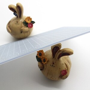 K&ouml;nnte beinhalten: Zwei Keramik-Hasenfiguren mit floralen Akzenten. Der obere Hase h&auml;lt einen Strau&szlig; gelber und roter Blumen. Der untere Hase h&auml;lt eine einzelne rote Blume. Eine transparente Kunststoff-Schneidematte ist auf den Hasen balanciert.
