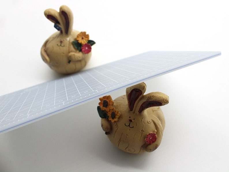 K&ouml;nnte beinhalten: Zwei Keramik-Hasenfiguren mit floralen Akzenten. Der obere Hase h&auml;lt einen Strau&szlig; gelber und roter Blumen. Der untere Hase h&auml;lt eine einzelne rote Blume. Eine transparente Kunststoff-Schneidematte ist auf den Hasen balanciert.