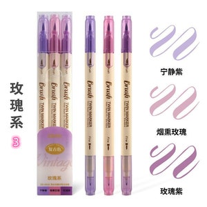 May include: A set of purple and pink twin-tip brush markers with the text "Brush TWIN MARKER" on the barrel. The packaging includes the text "Vintage" and Chinese characters. The image also shows examples of brush strokes in shades of purple.