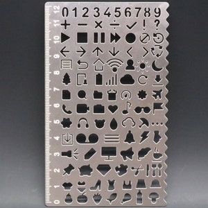 May include: A silver metal stencil with a ruler on the side and a variety of symbols and icons, including numbers, arrows, shapes, and musical notes.
