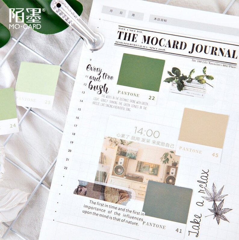 May include: A green and white journal with a grid pattern and stickers. The stickers feature a green color swatch with the text "PANTONE 22", a photo of a room with a speaker and plants, and a brown color swatch with the text "PANTONE 45". The journal also has the text "The Mocard Journal" and "The 100 Life Magazines 001/Plant".