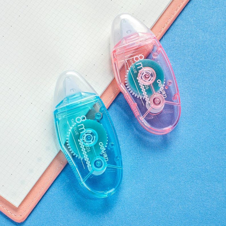 May include: Two clear plastic tape dispensers with blue and pink bodies. The dispensers have a small, clear, round tape roll inside. The tape dispensers have the text "8 mm 32B" and "deSIGN" printed on them.