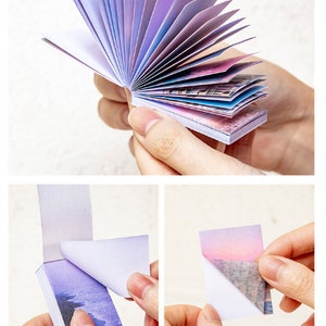 May include: A stack of colourful sticky notes with a gradient of purple, pink, and blue. The notes are folded in a fan shape and have a white backing. The notes have a variety of images printed on them, including a cityscape and a sunset.