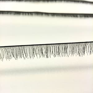 Op de afbeelding: Een close-up van een zwarte wimperstrip met een rij individuele wimpers. De wimpers zijn in een rechte lijn gerangschikt en gelijkmatig verdeeld.