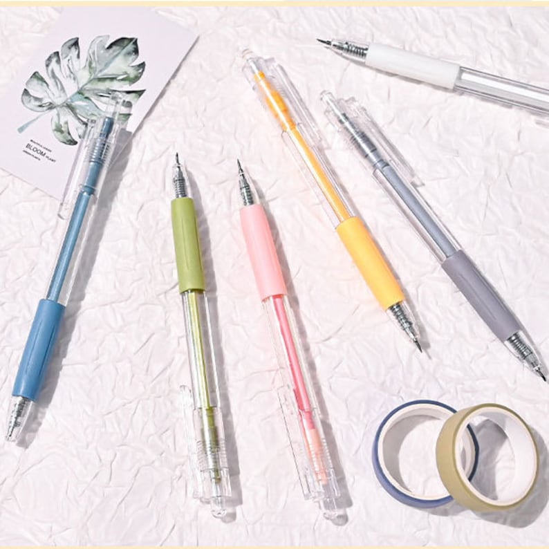 Puede incluir: Un conjunto de seis bol&iacute;grafos transparentes con insertos de tinta de colores. Los bol&iacute;grafos est&aacute;n dispuestos sobre una superficie blanca con un rollo de cinta washi. Los bol&iacute;grafos son azules, verdes, rosas, amarillos, grises y blancos.
