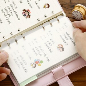 May include: A pink leather bound notebook with a clear plastic insert for holding loose papers. The notebook has six silver rings and a silver clasp. The notebook is open to a page with handwritten text and a cartoon illustration of a couple holding hands.