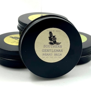 May include: Four black tins of beard balm are stacked on a white surface. The front tin has a label that reads "SOUTHERN GENTLEMAN BEARD BALM" and "net wt. 2oz (56.70g)". The other tins have labels with text and a silhouette.