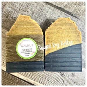 May include: Two rectangular bars of walnut scrub exfoliating soap. The top portion is light brown, the bottom is dark gray. One bar is wrapped with a brown paper label that reads "Walnut Scrub" and "net wt 5oz (142g)".