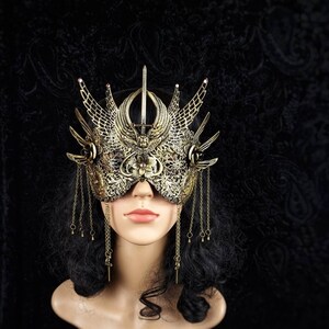 May include: A gold masquerade mask with intricate details and dangling chains. The mask is worn by a mannequin with black hair.