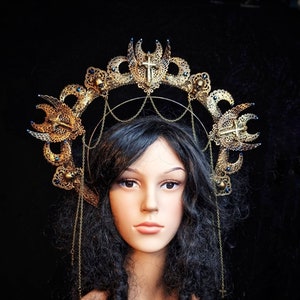 Könnte beinhalten: Eine goldene Krone mit einem Heiligenschein-Design, verziert mit filigranen Arbeiten und blauen Akzenten. Die Krone ist mit kleinen Kreuzen geschmückt und wird von einer Mannequin mit langen schwarzen Haaren getragen.