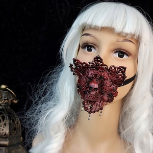 Könnte beinhalten: Eine rote Spitzenmaske mit Totenkopfdesign und herabhängenden Perlen. Die Maske wird von einer Puppe mit weißen Haaren getragen.