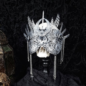May include: A silver skull crown with intricate details and dangling chains. The crown is displayed on a black velvet stand against a black background.