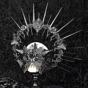 May include: A black skull with a crown of black butterflies and gold accents, surrounded by a halo of sharp black spikes. The skull is on a black pedestal against a black background.
