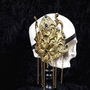 Könnte beinhalten: Ein aufwendiger, goldfarbener Kopfschmuck mit floralen und ornamentalen Details sowie hängenden Ketten. Er wird auf einem weißen Schädel vor dunklem Hintergrund präsentiert, was auf ein gotisches oder Fantasy-Thema hindeutet. Der Kopfschmuck ist wahrscheinlich aus Metall.