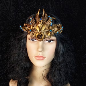 May include: A gold crown with a skull and rose design, accented with blue rhinestones. The crown is worn on a mannequin with long black hair.