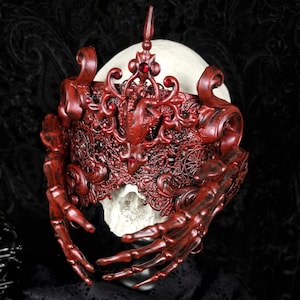 May include: A detailed red mask with a skeletal hand design. The mask has a crown and red jewels. The mask is set against a dark background, with black lace fabric in the foreground. The mask is an example of gothic art.