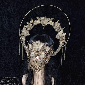May include: A gold-toned filigree mask and halo headpiece featuring intricate butterfly motifs, floral patterns, and delicate metal chains. The mask covers the upper face and nose, while the circular halo rises above the head with dangling accents.