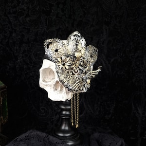 May include: A gold and black skull crown with intricate details and dangling chains. The crown is displayed on a white skull on a black lace background.