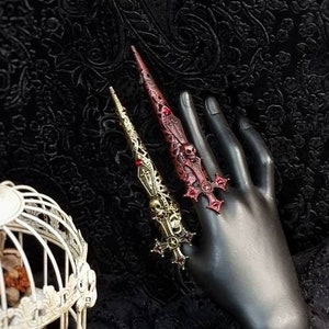 May include: Two long, pointed finger armor rings featuring intricate sword and skull motifs. One ring is finished in a metallic gold tone, while the other is a deep, dark red. Both pieces are adorned with small, embedded red crystals.