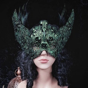 May include: A detailed, green mask with wing-like extensions and feather accents. The mask features intricate floral and ornamental designs. The mask is displayed on a mannequin head with dark curly hair, against a dark background.