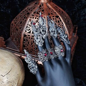 Könnte beinhalten: Eine schwarze Hand trägt einen Satz silberner Ringe mit roten Edelsteinen. Die Ringe sind verziert und weisen komplizierte Designs auf. Die Hand ruht auf einem schwarzen Stoff mit einem Schädel im Vordergrund.