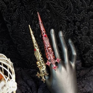 May include: Two long, pointed finger armor rings featuring intricate filigree metalwork. One ring is gold-toned with a skull and cross motif, while the other is dark red with a skull and cross design. Both pieces are adorned with small red accents.