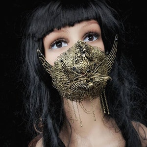 Könnte beinhalten: Eine goldfarbene Metallmaske mit filigranen Details und herabhängenden Ketten. Die Maske ist in Form eines Schmetterlings mit einem floralen Design gestaltet und wird von einer Mannequin mit schwarzen Haaren getragen.