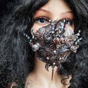 May include: A silver and copper masquerade mask with intricate details and rhinestones. The mask has a pointed, sharp piece extending from the bottom.