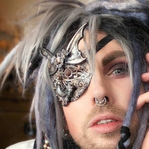 Könnte beinhalten: Eine Person mit grauen und blauen Dreadlocks, die eine silberne Augenklappe mit einem dekorativen Design trägt. Die Person trägt auch eine schwarze Krawatte, mehrere Armbänder und Ringe. Die Augenklappe hat ein dekoratives Design.