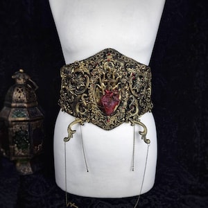 Könnte beinhalten: Verzierter, goldfarbener Gürtel mit zentralem Herzdesign. Der Gürtel weist aufwendige Schnörkel, rote Akzente und hängende Ketten mit kleinen Anhängern auf. Der Gürtel wird auf einer weißen Schaufensterpuppe vor dunklem Hintergrund präsentiert.