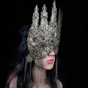 Puede incluir: Una m&aacute;scara de metal dorado para un baile de m&aacute;scaras con detalles intrincados y cadenas colgantes. La m&aacute;scara cubre los ojos y parte de la frente, con un dise&ntilde;o puntiagudo en forma de corona.