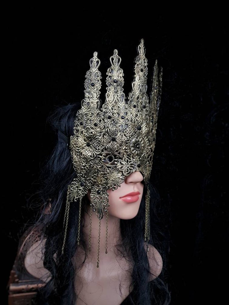 Puede incluir: Una m&aacute;scara de metal dorado para un baile de m&aacute;scaras con detalles intrincados y cadenas colgantes. La m&aacute;scara cubre los ojos y parte de la frente, con un dise&ntilde;o puntiagudo en forma de corona.