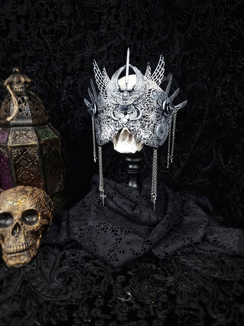 May include: A silver skull crown with intricate details and dangling chains. The crown is displayed on a black velvet stand against a black background.