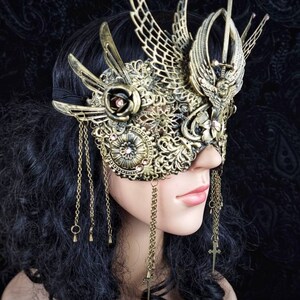 May include: A gold masquerade mask with intricate details, including a rose, wings, and a sword. The mask is adorned with rhinestones and has dangling chains.
