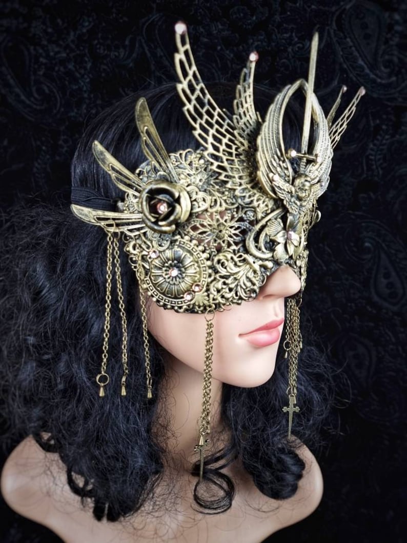 May include: A gold masquerade mask with intricate details, including a rose, wings, and a sword. The mask is adorned with rhinestones and has dangling chains.