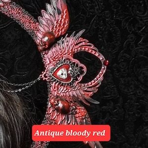 May include: A decorative headpiece featuring intricate red and black wings, small skulls, and a central heart emblem with an eye motif. The piece includes delicate chain accents and is labeled with the text Antique bloody red in a red box.