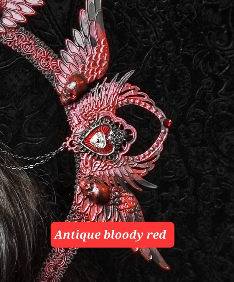 May include: A decorative headpiece featuring intricate red and black wings, small skulls, and a central heart emblem with an eye motif. The piece includes delicate chain accents and is labeled with the text Antique bloody red in a red box.