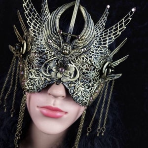 May include: A gold-coloured masquerade mask with intricate details, including a sword, wings, and floral accents. The mask is adorned with small pink rhinestones and has dangling gold chains.
