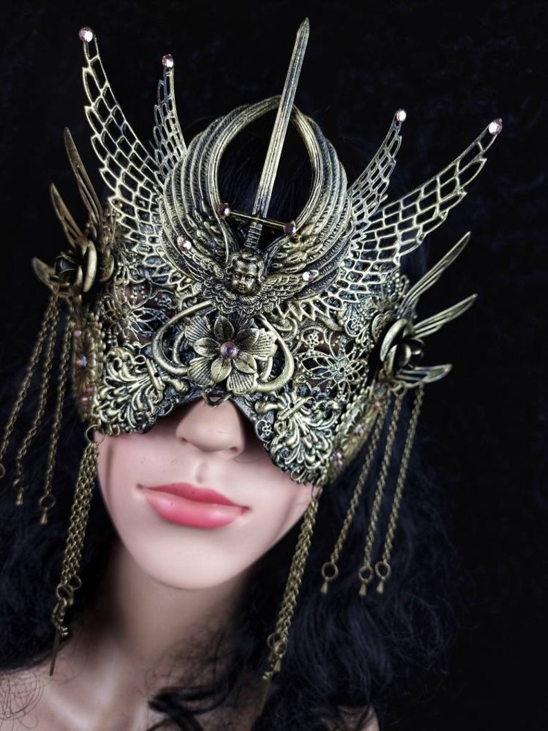 May include: A gold-coloured masquerade mask with intricate details, including a sword, wings, and floral accents. The mask is adorned with small pink rhinestones and has dangling gold chains.