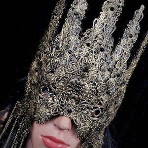 Puede incluir: Una m&aacute;scara de carnaval dorada y negra con detalles intrincados y cadenas colgantes. La m&aacute;scara tiene forma de corona y cubre los ojos.