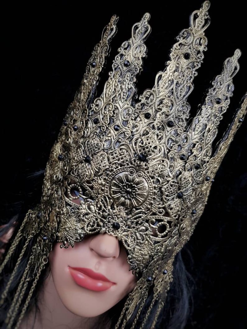 Puede incluir: Una m&aacute;scara de carnaval dorada y negra con detalles intrincados y cadenas colgantes. La m&aacute;scara tiene forma de corona y cubre los ojos.