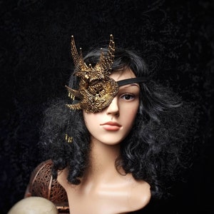 Könnte beinhalten: Ein goldfarbener Augenklappen mit filigranen Details und einem schwarzen Gummiband. Die Augenklappe wird von einer Mannequin mit langen, schwarzen, lockigen Haaren getragen.