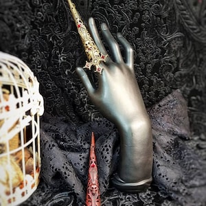 May include: A gold-toned metal finger armor ring featuring an elongated, tapered design with intricate filigree, skull motifs, and a cross-shaped base adorned with small red stones. The piece is displayed on a dark mannequin hand.