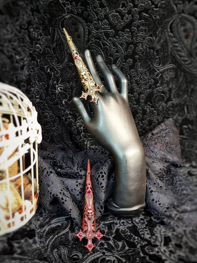 May include: A gold-toned metal finger armor ring featuring an elongated, tapered design with intricate filigree, skull motifs, and a cross-shaped base adorned with small red stones. The piece is displayed on a dark mannequin hand.
