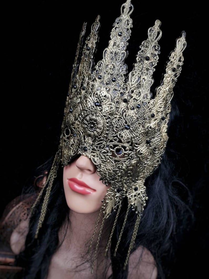 Puede incluir: Una m&aacute;scara de masquerade de oro en forma de corona con detalles negros y cadenas colgantes. La m&aacute;scara est&aacute; hecha de metal y tiene un dise&ntilde;o delicado e intrincado.