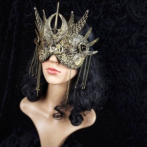 May include: A gold masquerade mask with intricate details and a crown-like design. The mask features a delicate, filigree pattern and dangling chains with small charms.