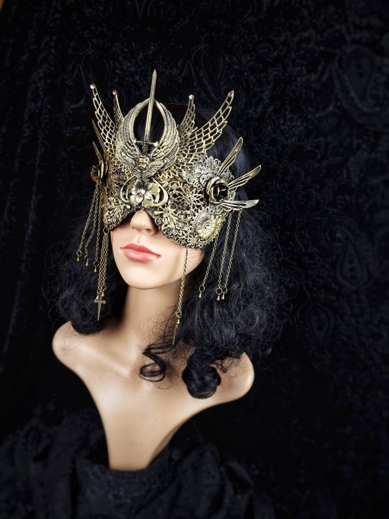 May include: A gold masquerade mask with intricate details and a crown-like design. The mask features a delicate, filigree pattern and dangling chains with small charms.